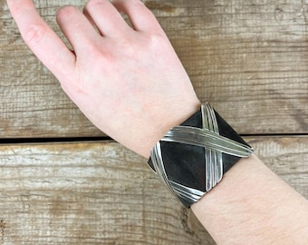 May include: A silver cuff bracelet with a black background and a woven design. The bracelet is worn on a person's wrist.