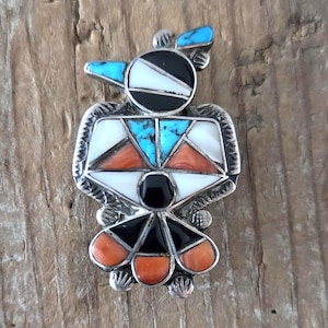 Op de afbeelding: Een zilveren broche in de vorm van een vogel, voorzien van inlegwerk van turkoois, witte schelp, zwarte onyx en oranje koraal. Het metalen frame heeft getextureerde randen, een geometrisch ontwerp, een kleine kuif op de kop en een waaiervormige staart.