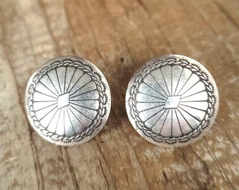 May include: Two silver-toned, round stud earrings. Each earring features a central square design, surrounded by a sunburst pattern and a beaded edge. The earrings are displayed on a wooden surface.