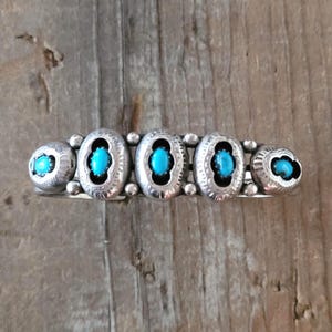 Puede incluir: Una pulsera de plata con cinco piedras ovaladas de turquesa engastadas en biseles ornamentados. La pulsera tiene un estilo vintage del suroeste, con una serie de pequeñas cuentas de plata que conectan las piedras más grandes. Las piedras de turquesa son de color azul vibrante.