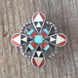 Può includere: Una spilla in argento con un cerchio centrale turchese circondato da quattro petali a forma di goccia e quattro segmenti triangolari. L'intarsio è composto da pietre rosse, nere, bianche e turchesi disposte in motivi geometrici in metallo.