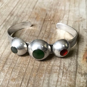May include: A silver bangle bracelet with three round, dome-shaped charms. Each charm has a different coloured stone inset: green, green, and red.