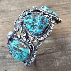 Puede incluir: Un brazalete de plata con tres grandes piedras de turquesa irregulares con patrones de matriz oscuros. El trabajo en metal incluye volutas intrincadas, motivos de hojas y bordes de cuerda trenzada alrededor de cada engaste.