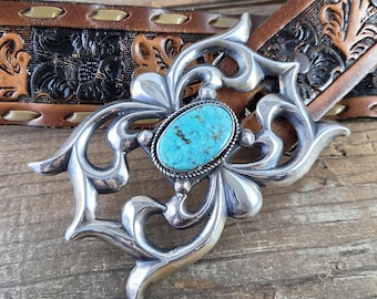 May include: A silver belt buckle featuring an oval turquoise stone set in the centre of an ornate, polished metal frame with scrolling details, attached to a brown leather belt with embossed floral patterns and stitched edges.