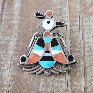 May include: A silver brooch featuring a stylized bird design with inlaid stone segments in black, white, turquoise, and orange. The metalwork displays textured details on the wings and base, with a central geometric pattern forming the bird's body.