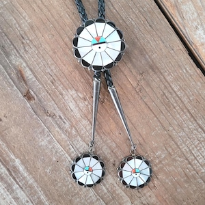 May include: A bolo tie featuring a large circular centerpiece and two smaller matching circular pendants at the ends of a braided black leather cord. The circular pieces are inlaid with white, black, turquoise, and coral segments in a sunburst pattern.