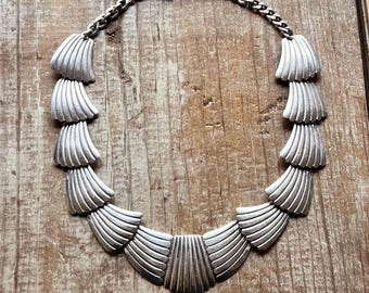 May include: A silver necklace with a geometric design. The necklace is made of a series of overlapping, fan-shaped pieces that create a cascading effect.