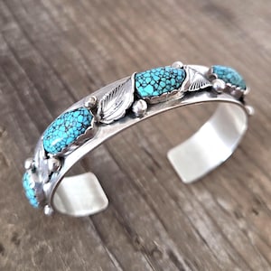 May include: A silver cuff bracelet featuring three oval turquoise stones with a dark matrix pattern. The bracelet is decorated with polished silver leaf motifs and small silver spheres placed between the stones along the band.