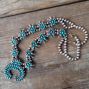 Puede incluir: Un conjunto de collar y pulsera de plata con detalles de piedras de color turquesa. El collar presenta un diseño en forma de media luna con grupos de piedras turquesas. La pulsera tiene un diseño simple de cuentas con un cierre de gancho.