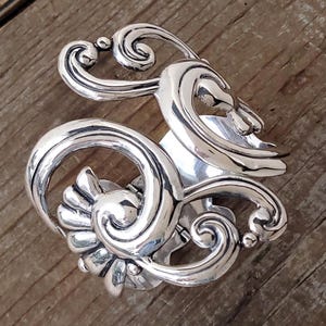 May include: A silver-toned, ornate brooch with a swirling, Art Nouveau design. The brooch features flowing lines, curls, and a floral-like element. The metal has a polished finish, reflecting light. The brooch is set against a wooden surface.