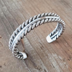 Puede incluir: Una pulsera de plata con diseño de cuerda trenzada. La pulsera tiene un acabado pulido y está abierta en la parte inferior. El metal es de color plata brillante. La pulsera tiene aproximadamente 6,35 cm de diámetro.