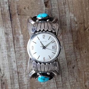 Op de afbeelding: Een zilverkleurig Nivia horloge met een witte wijzerplaat, zwarte cijfers en een datumvenster. Het horloge is gezet in een brede manchetarmband met turkooizen stenen en decoratieve bloemelementen.