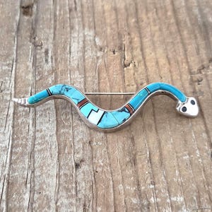 May include: A silver snake-shaped brooch featuring inlaid segments of bright blue turquoise, white stone, and thin dark brown accents. The serpent body has a curved, undulating form with two small black circular eyes on the head and a pin back.