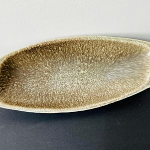 May include: A ceramic serving dish with an unusual, elongated shape. The interior has a speckled brown and tan pattern, while the rim is a solid, light beige. Suitable for serving appetizers or small dishes.