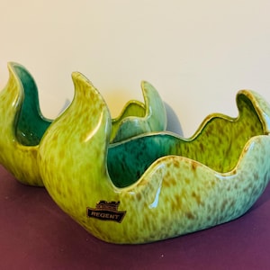 May include: Two ceramic bowls with a wavy, organic rim design. The exterior and interior feature a mottled lime green and brown glaze with a glossy finish. A black label with the text REGENT is affixed to the front of the larger bowl.