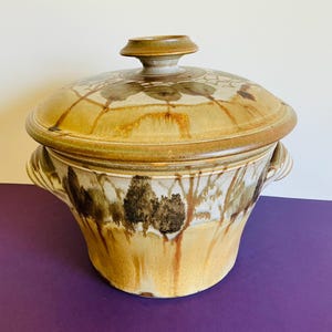 May include: A round ceramic lidded casserole dish featuring a tan and brown drip glaze. The body displays a dark brown landscape pattern of trees, while the lid has a matching floral motif and a central knob handle for easy lifting.
