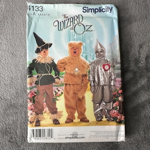 Puede incluir: Patrón de costura Simplicity para disfraces de El Mago de Oz. El patrón incluye instrucciones para los disfraces del Espantapájaros, el León Cobarde y el Hombre de Hojalata. El paquete es blanco y azul con imágenes de los disfraces y el texto "The Wizard of Oz".