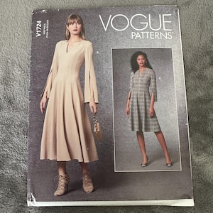 May include: Vogue Patterns V1724 sewing pattern featuring two dress designs. The dress on the left is a beige midi dress with long sleeves and slit details. The dress on the right is a gray plaid midi dress with elbow-length sleeves and slit details.
