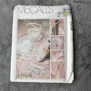 以下が含まれることがあります： ウェディングアクセサリーが描かれた、McCall'sのソーイングパターン封筒。ハート型の枕、ハンドバッグ、ガーター、バラの靴、手袋、ヘッドピースが写っています。「Alicyn Exclusives」の文字が見えます。