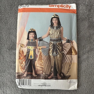 May include: Simplicity sewing pattern 2329 for Cleopatra costumes. The pattern features two costumes with gold, black, and blue accents. The costumes include headbands, armbands, and flowing skirts. The pattern is for sizes HH 3,4,5,6.
