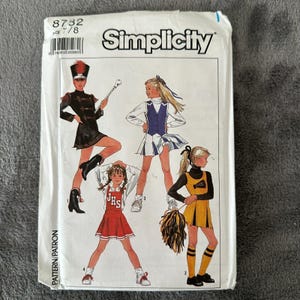 May include: A Simplicity sewing pattern envelope featuring illustrations of children in various costumes. The costumes include a marching band uniform, cheerleading outfits, and a school uniform. The envelope has the number 8732 and the Simplicity logo.
