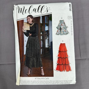 May include: A McCall's M8150 sewing pattern envelope featuring a tiered skirt design. The front shows a person wearing a black long-sleeved top and a dark, shimmering tiered skirt. Illustrations of skirt variations A and C are printed on the right.