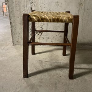 K&ouml;nnte beinhalten: Ein kleiner, quadratischer Holzschemel mit einem Sitz aus geflochtenem Stroh. Der Hocker hat vier dunkelbraune Beine und horizontale Streben. Der Sitz ist hellbeige und das Holz hat eine nat&uuml;rliche Oberfl&auml;che. Der Hocker ist ein einfaches, funktionelles M&ouml;belst&uuml;ck.