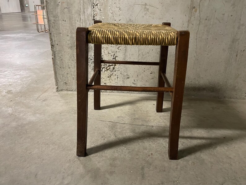 K&ouml;nnte beinhalten: Ein kleiner, quadratischer Holzschemel mit einem Sitz aus geflochtenem Stroh. Der Hocker hat vier dunkelbraune Beine und horizontale Streben. Der Sitz ist hellbeige und das Holz hat eine nat&uuml;rliche Oberfl&auml;che. Der Hocker ist ein einfaches, funktionelles M&ouml;belst&uuml;ck.