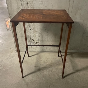 Peut inclure: Une table d'appoint rectangulaire et haute en bois, dotée d'une finition brun foncé et d'un motif géométrique en placage sur le plateau. La table repose sur quatre pieds fins et droits reliés par de fines barres de support horizontales.