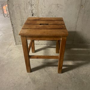 May include: A small wooden stool with a rectangular seat and four legs. The seat has a rectangular opening in the centre. The wood has a warm, brown tone and visible grain. The stool is sturdy and simple in design.