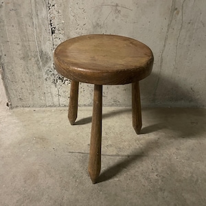 Op de afbeelding: Een kleine, ronde houten kruk met een gladde, cirkelvormige zitting en drie taps toelopende poten. De kruk is gemaakt van natuurlijk hout met een zichtbare nerf en een middenbruine afwerking, rustend op drie puntige voeten.