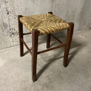 K&ouml;nnte beinhalten: Kleiner, quadratischer Hocker mit einer Sitzfl&auml;che aus geflochtenem Stroh und dunkelbraunen Holzbeinen. Der Sitz hat eine hellbraune Farbe mit einem gewebten Muster. Die Beine sind stabil und der Hocker scheint vintage zu sein.