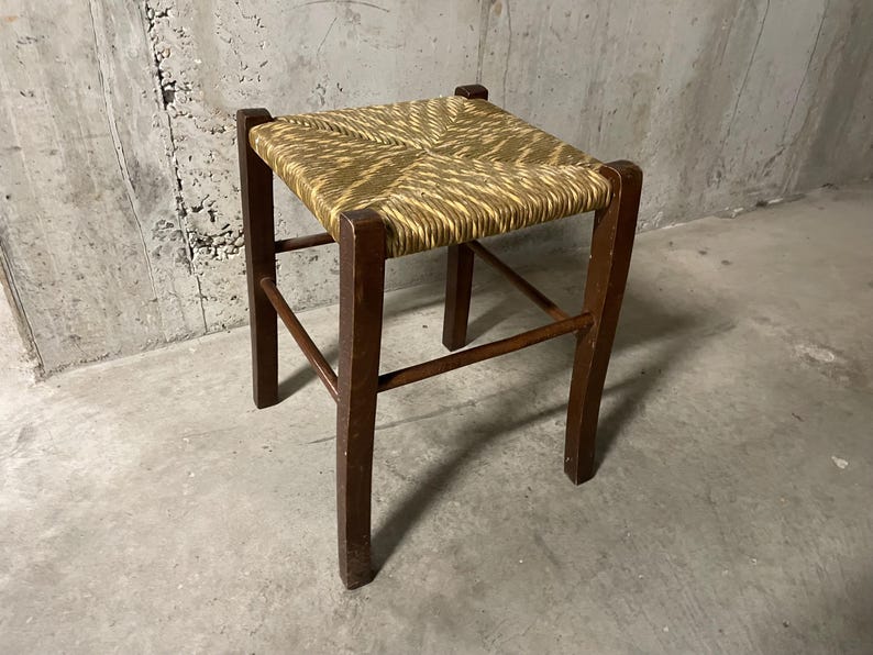 K&ouml;nnte beinhalten: Kleiner, quadratischer Hocker mit einer Sitzfl&auml;che aus geflochtenem Stroh und dunkelbraunen Holzbeinen. Der Sitz hat eine hellbraune Farbe mit einem gewebten Muster. Die Beine sind stabil und der Hocker scheint vintage zu sein.
