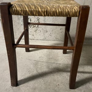 K&ouml;nnte beinhalten: Hocker aus Holz mit gewebter Strohsitzfl&auml;che. Die Beine sind dunkelbraun und rechteckig, verbunden durch horizontale und abgewinkelte Streben. Der Sitz hat eine hellbraune Farbe.