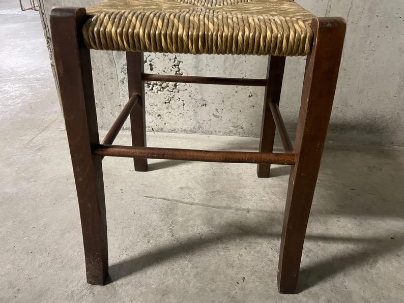 K&ouml;nnte beinhalten: Hocker aus Holz mit gewebter Strohsitzfl&auml;che. Die Beine sind dunkelbraun und rechteckig, verbunden durch horizontale und abgewinkelte Streben. Der Sitz hat eine hellbraune Farbe.