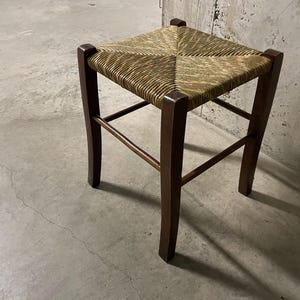 K&ouml;nnte beinhalten: Quadratischer Holzschemel mit gewebter Strohsitzfl&auml;che. Die dunkelbraunen Beine sind durch horizontale Streben verbunden. Der Hocker ist ein schlichtes, rustikales Design, das f&uuml;r verschiedene Einrichtungsstile geeignet ist. Der Sitz ist ca. 30 cm x 30 cm gro&szlig;.