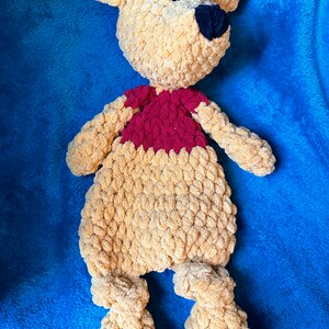 May include: A crocheted stuffed bear toy with a brown body and a red shirt. The bear is lying on a blue blanket.