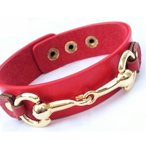 May include: Red leather bracelet with a gold horse bit buckle. The bracelet has four decorative holes on the band.