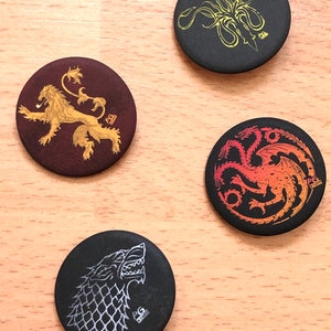 Può includere: Quattro bottoni rotondi neri e bianchi con diversi stemmi delle case di Game of Thrones. Gli stemmi sono un leone, un drago, un lupo e un kraken. Ogni bottone ha il testo "PG Stuff" in basso.