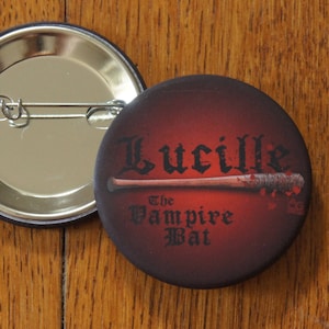 Peut inclure: Un bouton rouge et noir avec le texte "Lucille The Vampire Bat" et une batte de baseball rouge et brune avec des éclaboussures de sang.