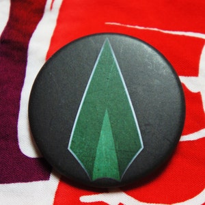 May include: A circular, black badge featuring a green arrow design. The arrow incorporates various shades of green and white. The badge is displayed on a vibrant, patterned fabric.