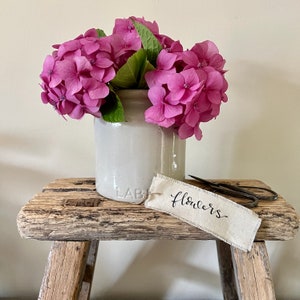 May include: A weathered wooden stool supports a cream-coloured ceramic vase overflowing with pink hydrangea blooms and green foliage. A tag reading "Flowers" and a pair of scissors are also on the stool.
