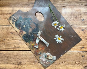 May include: A well-used wooden artist's palette with dried paint and three paint tubes. White daisies with yellow centres are arranged on the palette. The palette has a thumb hole and is on a wooden surface.