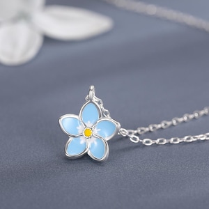 May include: A silver necklace with a light blue and yellow flower pendant. The flower pendant has five petals and a yellow centre. The necklace is shown on a grey surface with white flower accents.