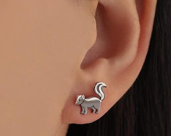 May include: A close-up of a silver skunk earring on a person's ear. The earring is a small, detailed representation of a skunk with a curved tail. The skunk is facing to the left.