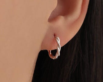 May include: A close-up of a silver hoop earring with a twisted design. The earring is worn on an ear, with the focus on the jewelry. The earring is a small, delicate piece of jewelry.