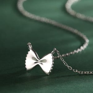 Pode incluir: Um colar prateado com um pendente em forma de massa farfalle. O pendente apresenta detalhes de nervuras e uma forma de laço. A delicada corrente pende sobre um fundo verde escuro. O pendente tem cerca de 2 cm de largura.