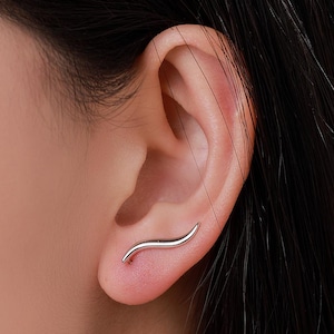 Könnte beinhalten: Ein silberner Ohrkletterer-Ohrring in einem geschwungenen, wellenartigen Design. Der Ohrring wird am Ohr getragen, wobei die Kurve der natürlichen Form des Ohrs folgt. Der Schmuck ist schlicht und elegant.