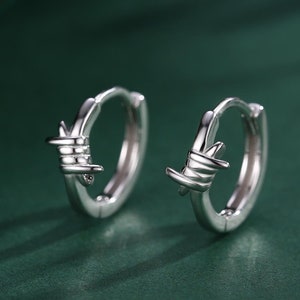 May include: A pair of silver hoop earrings with a barbed wire design. The earrings are on a green surface.