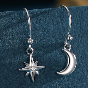 May include: Silver drop earrings with celestial motifs. One earring features a starburst with a central gem, the other a crescent moon. The earrings hang from delicate hooks, adding a touch of elegance to the design.